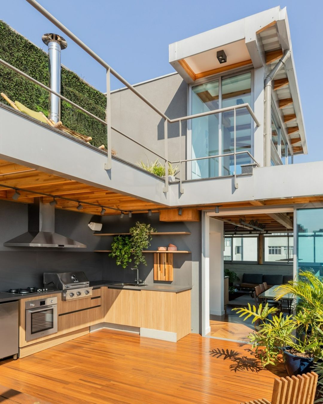 Área gourmet externa integrada à área social da casa, com churrasqueira, madeira aparente e portas de vidro que conectam os ambientes.