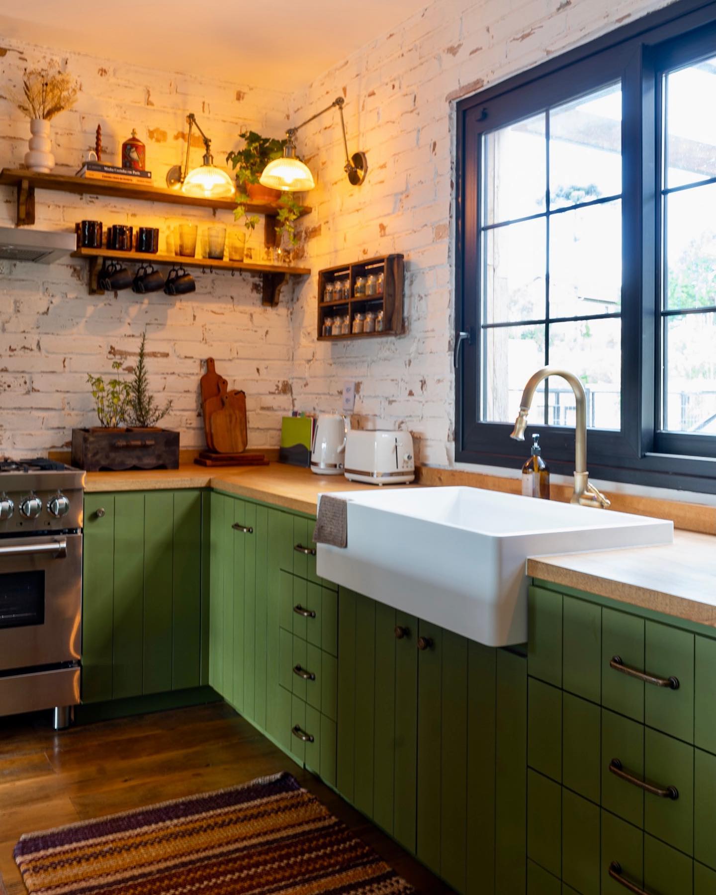 Cozinha estilo farmhouse com armários verdes, parede de tijolinhos brancos e janela preta.