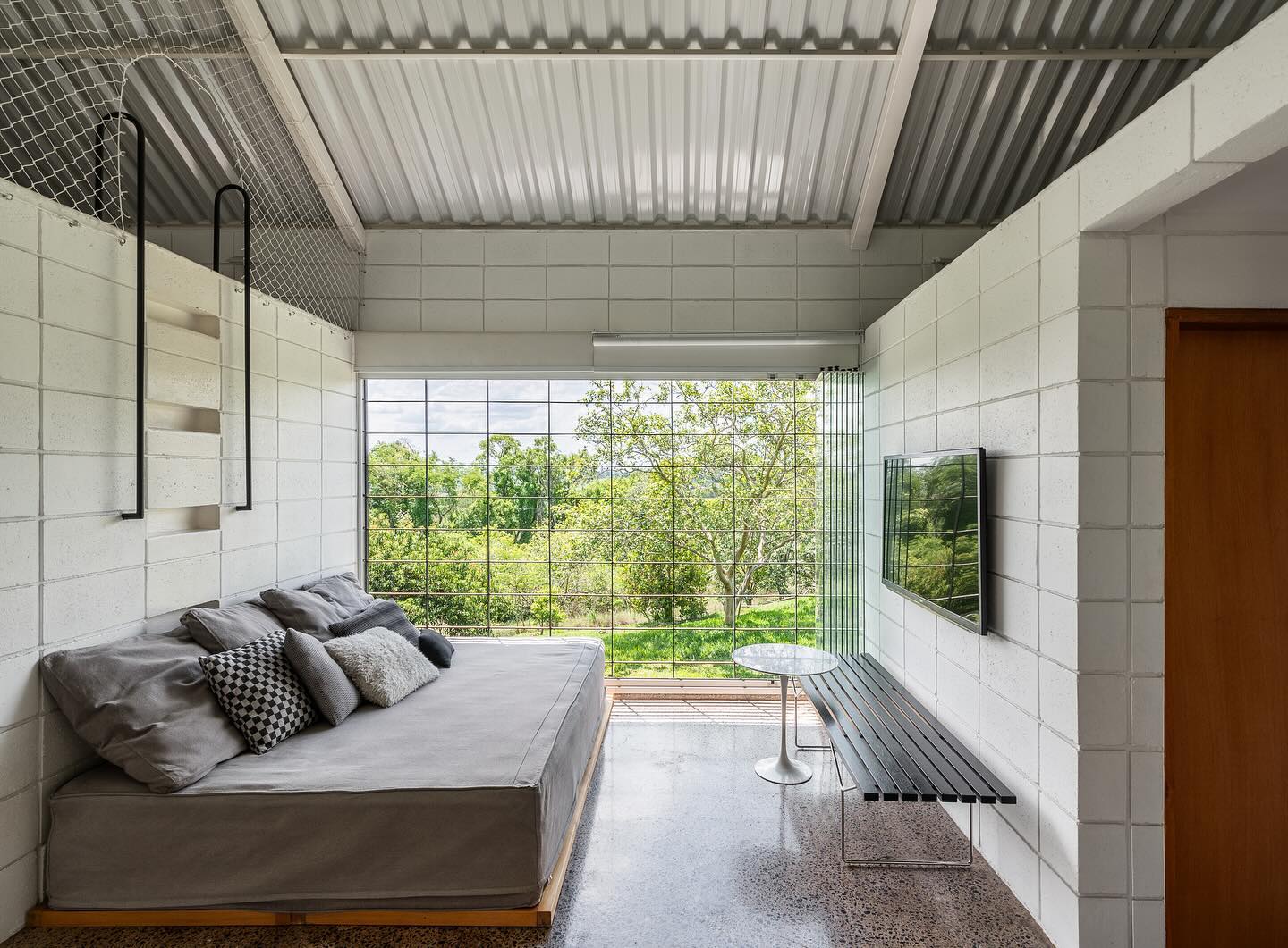 Vista da sala de estar da casa, composta por sofá, televisão suspensa na parede e banco de metal horizontal.