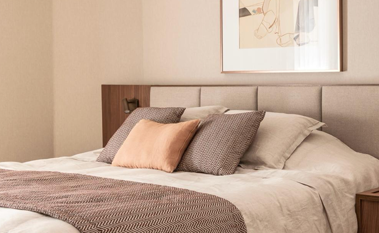 Quarto em tons greige com cabeceira estofada, roupa de cama neutra e detalhes em madeira.