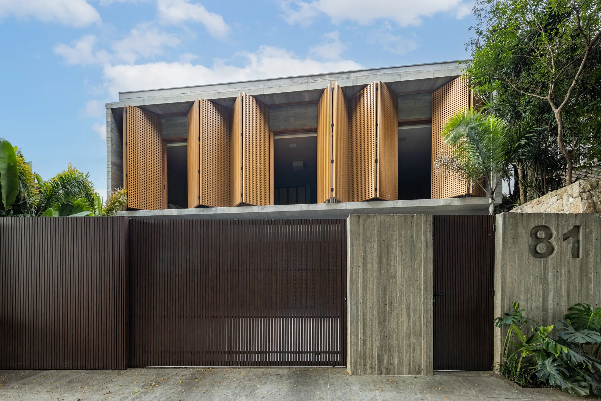 Fachada de casa em Moema com muxarabi, concreto aparente, madeira freijó e entrada voltada para praça arborizada.