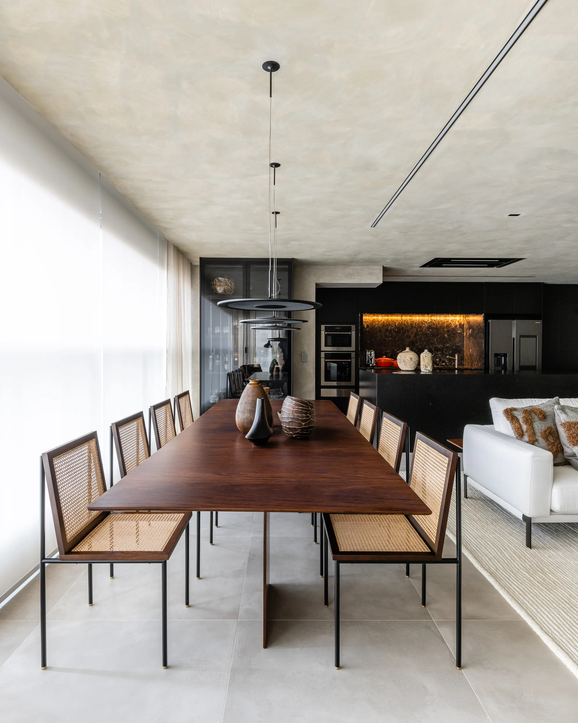 Sala de jantar com mesa de madeira escura, cadeiras de palhinha e pendentes circulares.
