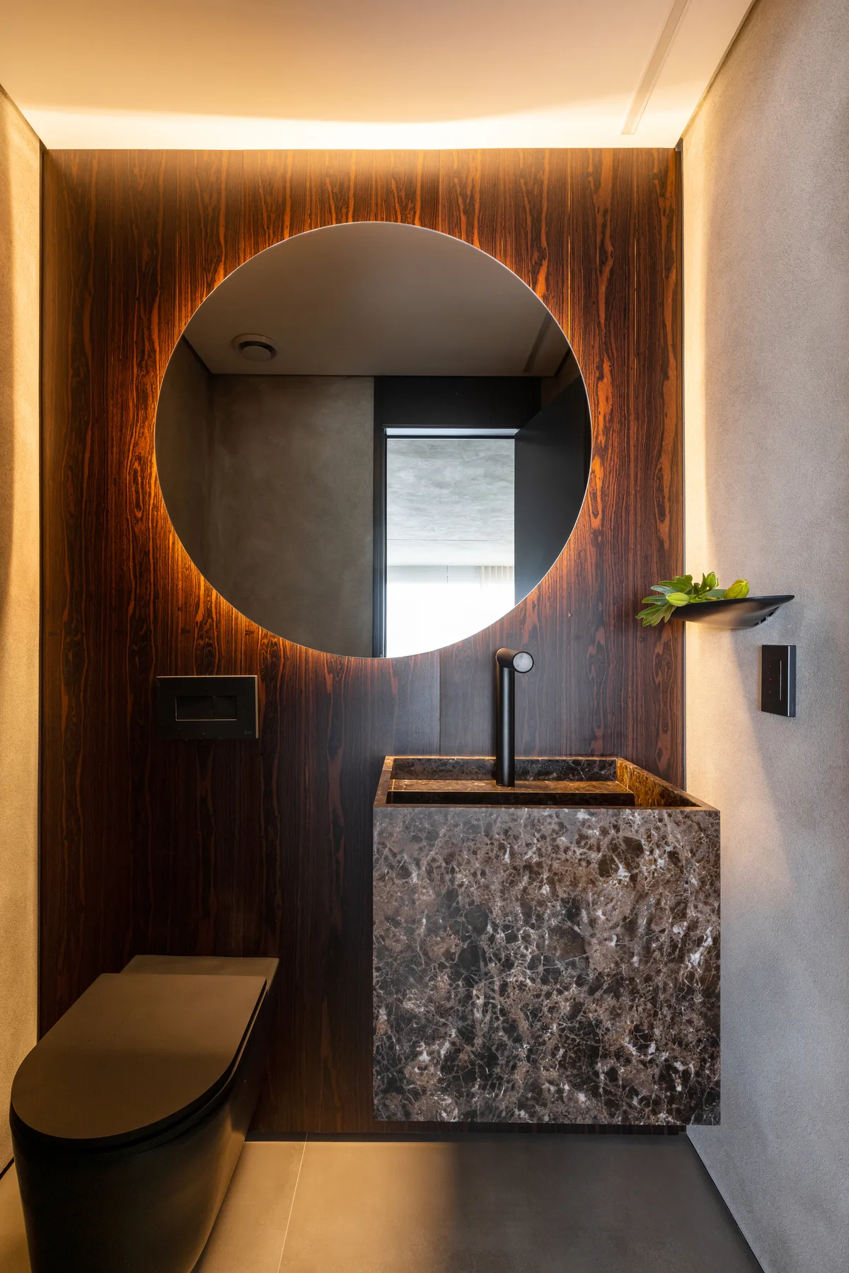 Lavabo revestido em madeira escura, com cuba esculpida em pedra marrom e espelho redondo iluminado.