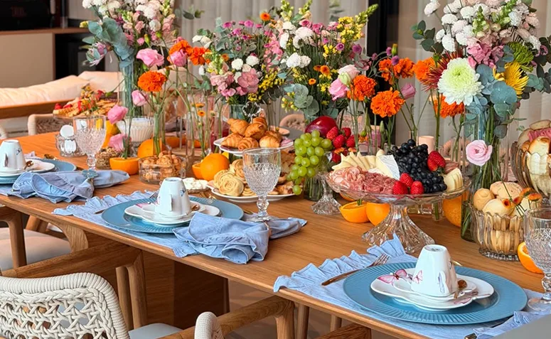 Mesa compartilhada de brunch com lugares montados, frutas, frios, pães e arranjos de flores