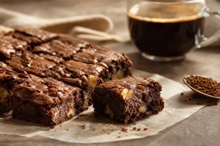 Quadrados de brownie de chocolate com nozes e café ao fundo.