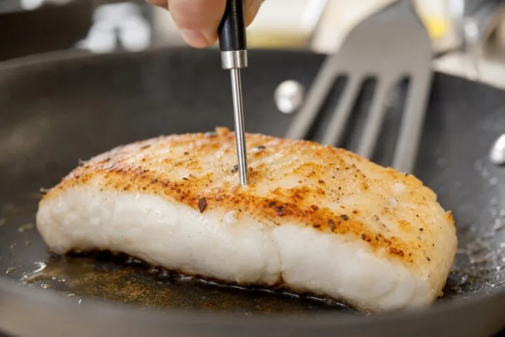 Termômetro culinário verificando o centro de um filé de peixe dourado na frigideira.