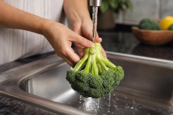 Mãos lavando uma cabeça inteira de brócolis com os floretes virados para baixo.