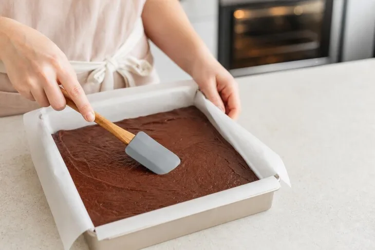 Massa de brownie sendo nivelada em forma quadrada de metal forrada.