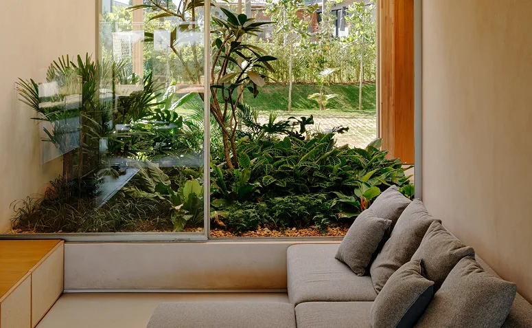 Jardim interno compacto com plantas tropicais e diferentes folhagens, integrado à sala por uma ampla janela de vidro.